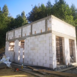 WOJCIECH MAMAK - Wznoszenie ścian parterowego budynku z białych bloczków, widoczne zbrojenia pionowe, w tle drzewa, na pierwszym planie paleta z bloczkami i piła spalinowa.