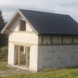 WOJCIECH MAMAK - Surowy budynek z białych bloczków z czarnym dachem z blachodachówki, widoczne niezabezpieczone otwory okienne i drzwiowe, drabina oparta o ścianę boczną.