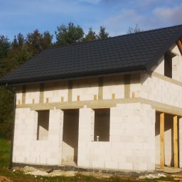 WOJCIECH MAMAK - Surowy stan domu z betonu komórkowego z zamontowaną czarną blachodachówką, widoczne drewniane elementy konstrukcyjne okien i drzwi, oraz fragment orynnowania.