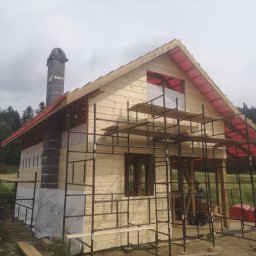 WOJCIECH MAMAK - Wznoszony drewniany dom z kominem z bloków, otoczony rusztowaniem, widoczne membrany dachowe i folie izolacyjne na ścianach, w tle zieleń drzew i pochmurne niebo.