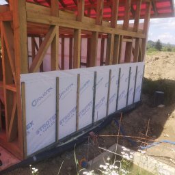 WOJCIECH MAMAK - Wznoszenie konstrukcji szkieletowej domu z widoczną membraną izolacyjną Foliatex Strotex Toples na ścianach, drewniane belki konstrukcyjne i widok na krajobraz w tle.