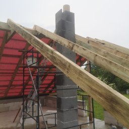 WOJCIECH MAMAK - Wznoszony komin z szarych bloczków betonowych na tle konstrukcji dachu z drewnianych belek i czerwonej membrany, widoczne rusztowanie i drabina.