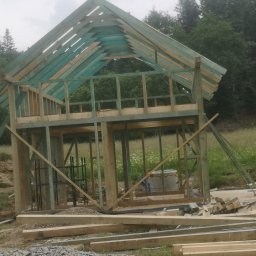 WOJCIECH MAMAK - Wznoszenie dwukondygnacyjnej konstrukcji drewnianej z elementami pomalowanymi na zielono, widoczne rusztowania i materiały budowlane na tle łąki i lasu.