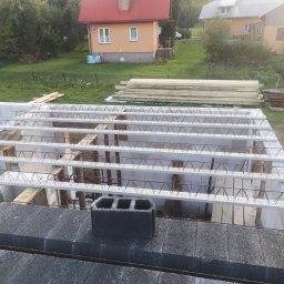 WOJCIECH MAMAK - Wiązanie zbrojenia stropu Teriva na budowie, widoczne bloczki betonowe, stalowe pręty i pustaki szalunkowe, w tle domy w zabudowie jednorodzinnej.
