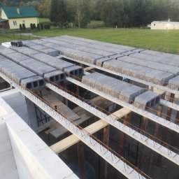 WOJCIECH MAMAK - Wieniec stropowy z betonowych belek i pustaków szalunkowych, widoczne zbrojenie, ściany z betonu komórkowego, w tle zabudowa jednorodzinna i zieleń.