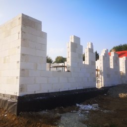 WOJCIECH MAMAK - Wznoszenie ścian z białych bloczków betonu komórkowego na fundamencie z izolacją przeciwwilgociową, widoczny fragment dachu domu w tle.
