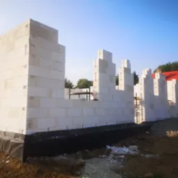 Wznoszenie ścian z białych bloczków betonu komórkowego na fundamencie z izolacją przeciwwilgociową, widoczny fragment dachu domu w tle.