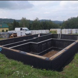 WOJCIECH MAMAK - Wykonane fundamenty domu z izolacją przeciwwilgociową, widoczne bloczki fundamentowe i folia kubełkowa, w tle zaparkowany biały bus i ułożone palety z bloczkami Solbet, krajobraz pagórkowaty.