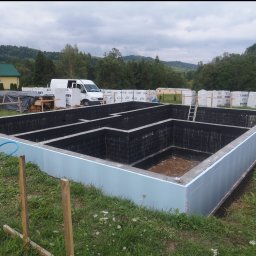 WOJCIECH MAMAK - Zalane fundamenty domu z hydroizolacją i ociepleniem, na tle zieleni i zabudowań, widoczny samochód dostawczy i materiały budowlane.