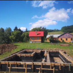 Zalany fundament z drewnianym szalunkiem na tle wiejskich domów i pagórkowatego krajobrazu w słoneczny dzień.