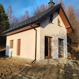 WOJCIECH MAMAK - Niewielki, parterowy dom z ciemną dachówką i drewnianą stolarką okienną, otoczony naturalnym krajobrazem w jesiennej scenerii. Widok z boku, w słoneczny dzień.