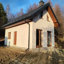 Niewielki, parterowy dom z ciemną dachówką i drewnianą stolarką okienną, otoczony naturalnym krajobrazem w jesiennej scenerii. Widok z boku, w słoneczny dzień.