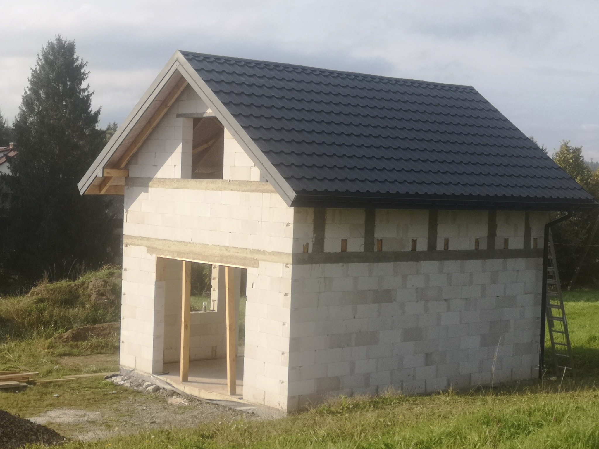 Surowy budynek z białych bloczków z czarnym dachem z blachodachówki, widoczne niezabezpieczone otwory okienne i drzwiowe, drabina oparta o ścianę boczną.