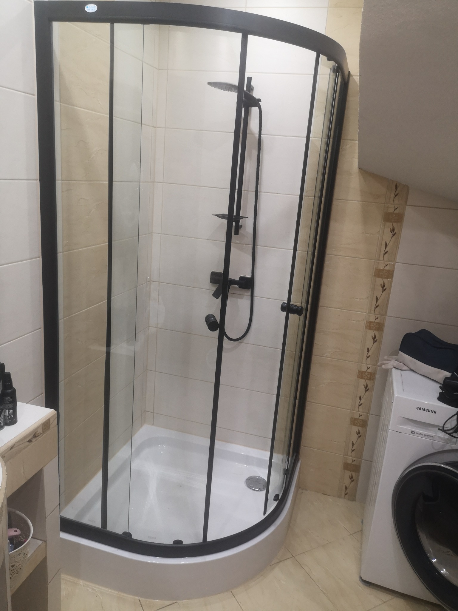 Kabina prysznicowa narożna z czarnymi profilami i deszczownicą, zamontowana w łazience z beżowymi płytkami, obok pralka Samsung.