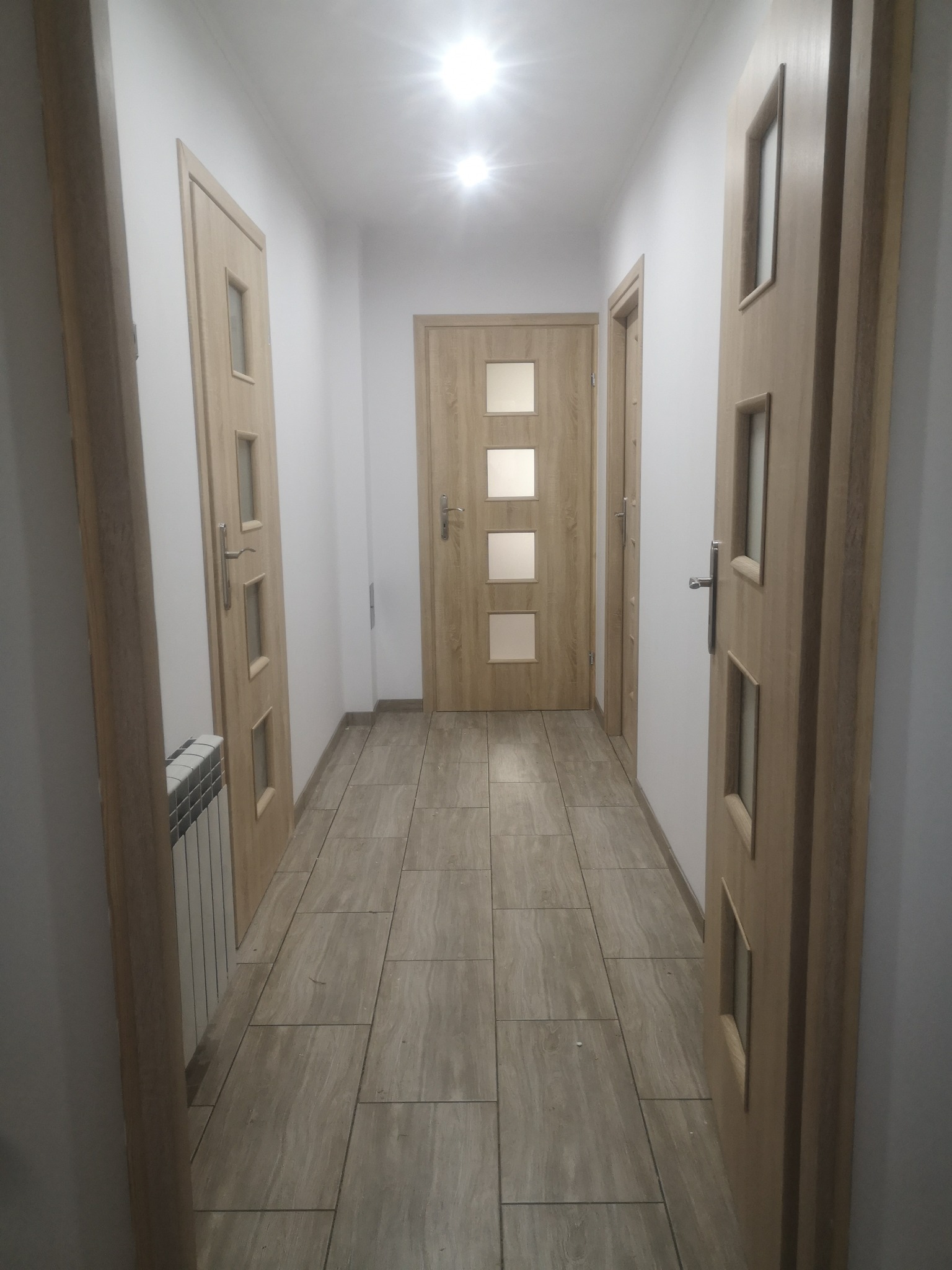 Wnętrze korytarza z trzema drewnianymi drzwiami z przeszkleniami, wykończonego płytkami podłogowymi w odcieniach beżu i szarości, widoczny grzejnik.