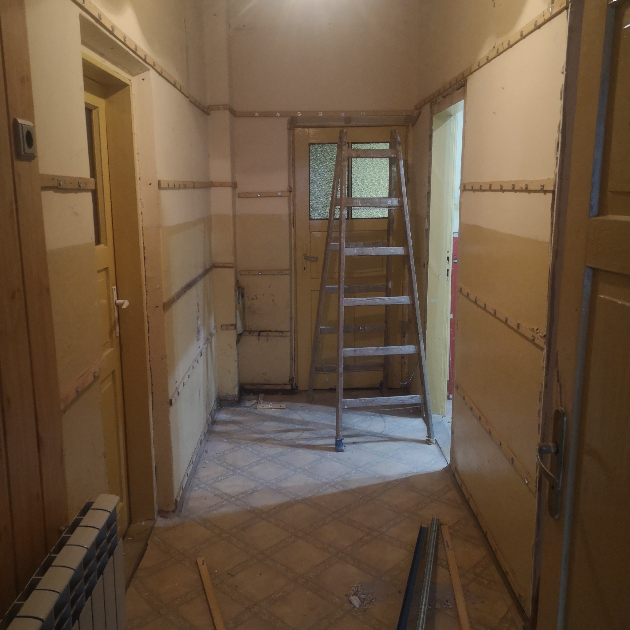 Korytarz w trakcie przygotowania do remontu: ściany z listwami, stara drabina oparta o drzwi, grzejnik i fragment podłogi z wzorem rombów.