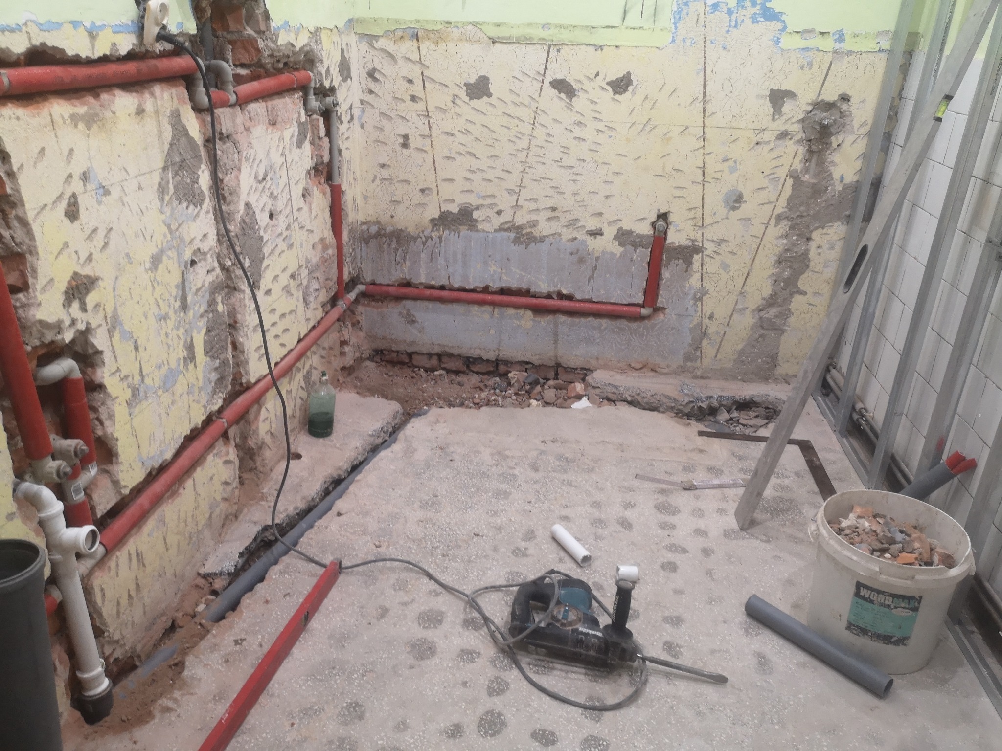 Wnętrze w trakcie remontu: odsłonięte instalacje wodne w czerwonych rurach na tle starej, zniszczonej ściany, narzędzia budowlane i wiadro z gruzem na betonowej posadzce.