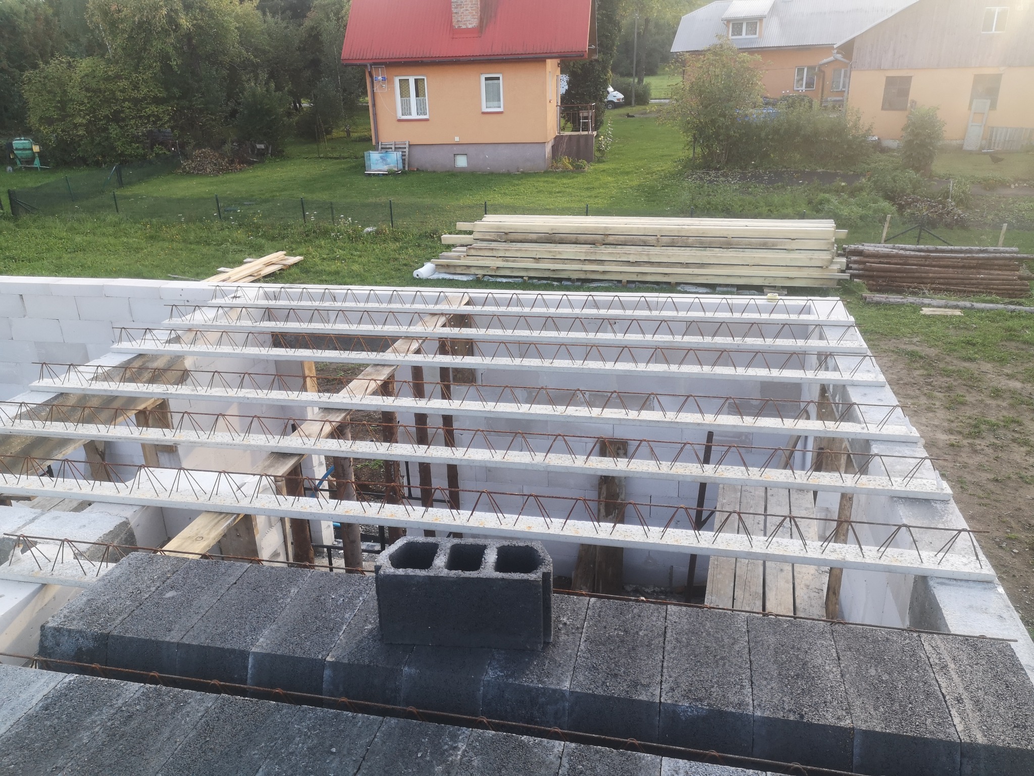 Wiązanie zbrojenia stropu Teriva na budowie, widoczne bloczki betonowe, stalowe pręty i pustaki szalunkowe, w tle domy w zabudowie jednorodzinnej.