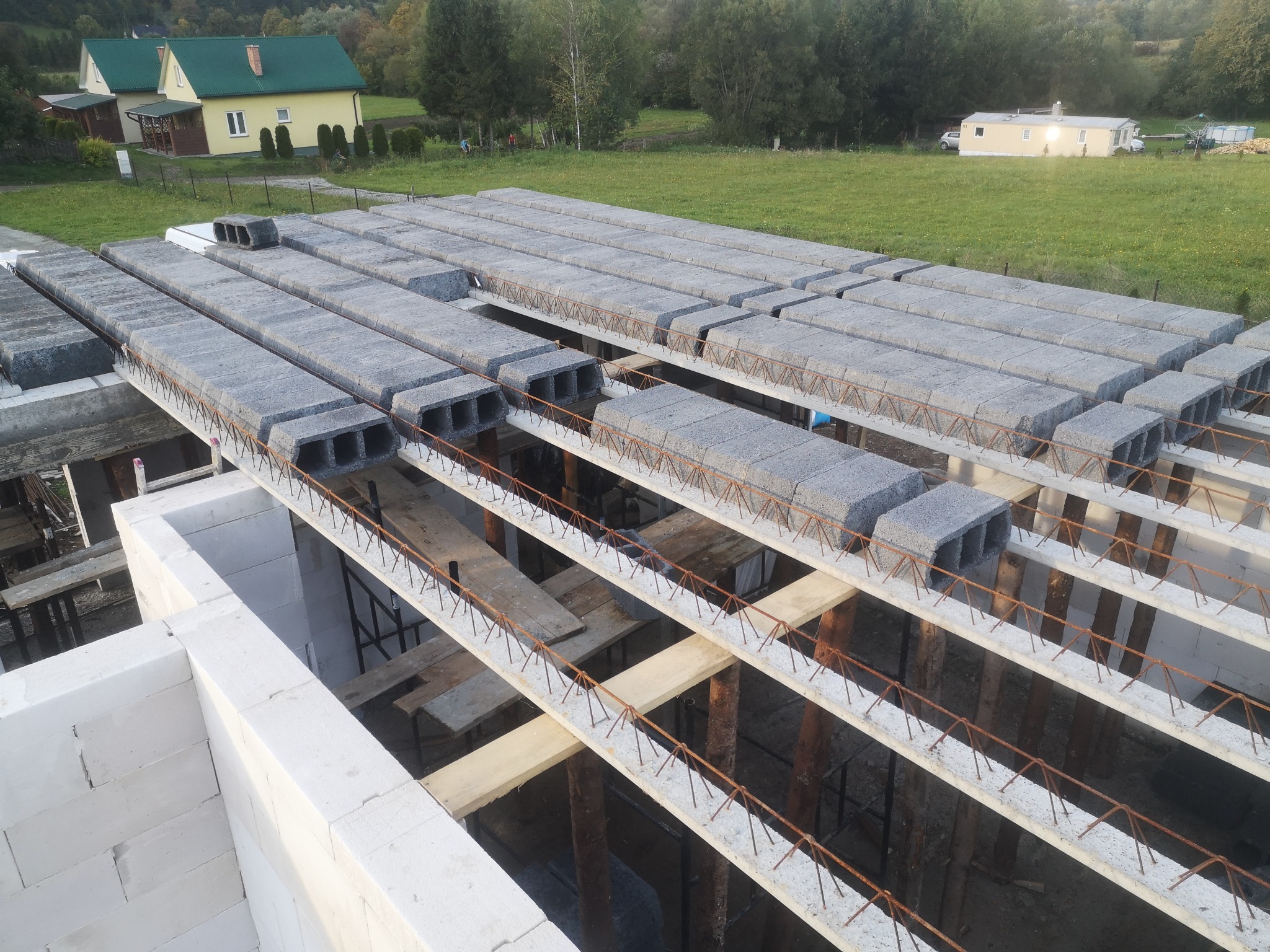 Wieniec stropowy z betonowych belek i pustaków szalunkowych, widoczne zbrojenie, ściany z betonu komórkowego, w tle zabudowa jednorodzinna i zieleń.