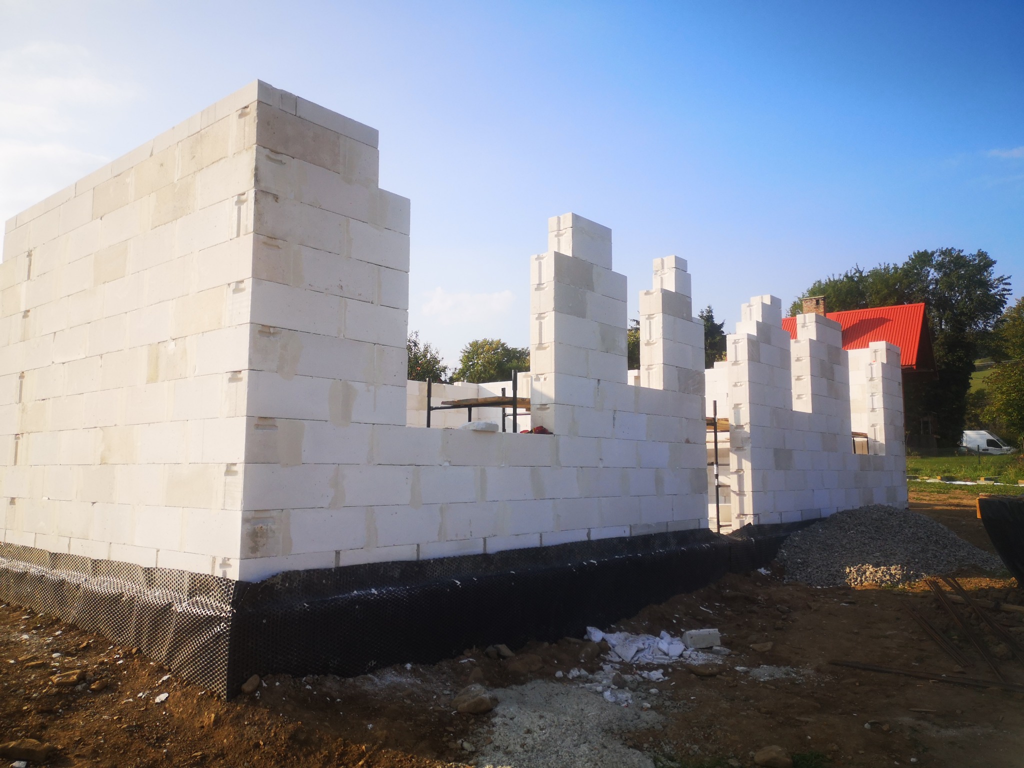 Wznoszenie ścian z białych bloczków betonu komórkowego na fundamencie z izolacją przeciwwilgociową, widoczny fragment dachu domu w tle.