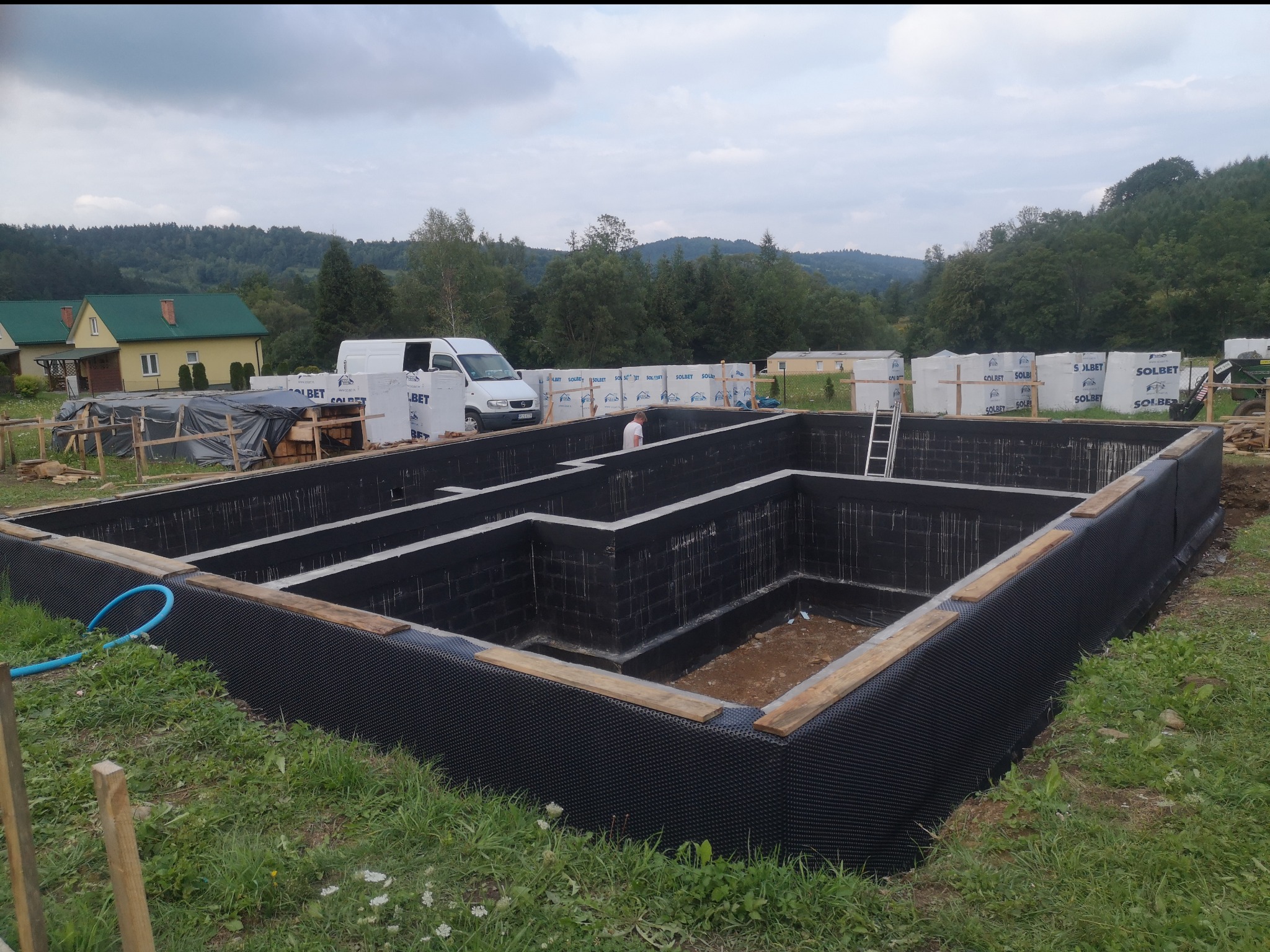 Wykonane fundamenty domu z izolacją przeciwwilgociową, widoczne bloczki fundamentowe i folia kubełkowa, w tle zaparkowany biały bus i ułożone palety z bloczkami Solbet, krajobraz pagórkowaty.