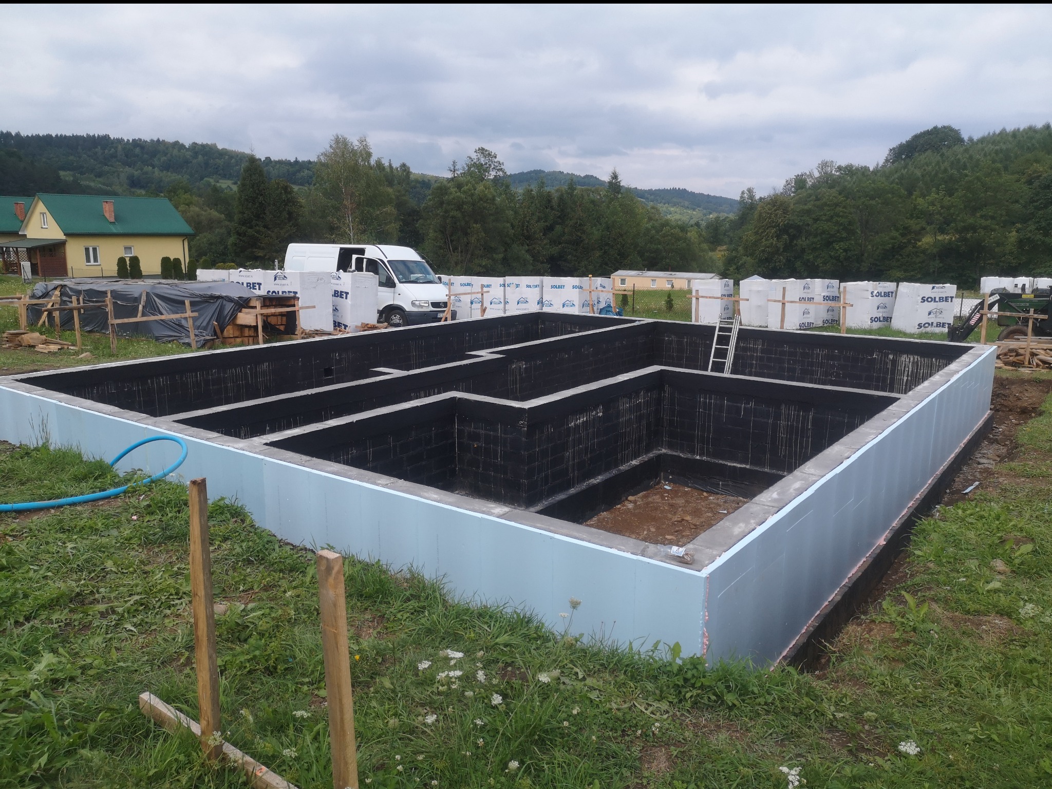 Zalane fundamenty domu z hydroizolacją i ociepleniem, na tle zieleni i zabudowań, widoczny samochód dostawczy i materiały budowlane.