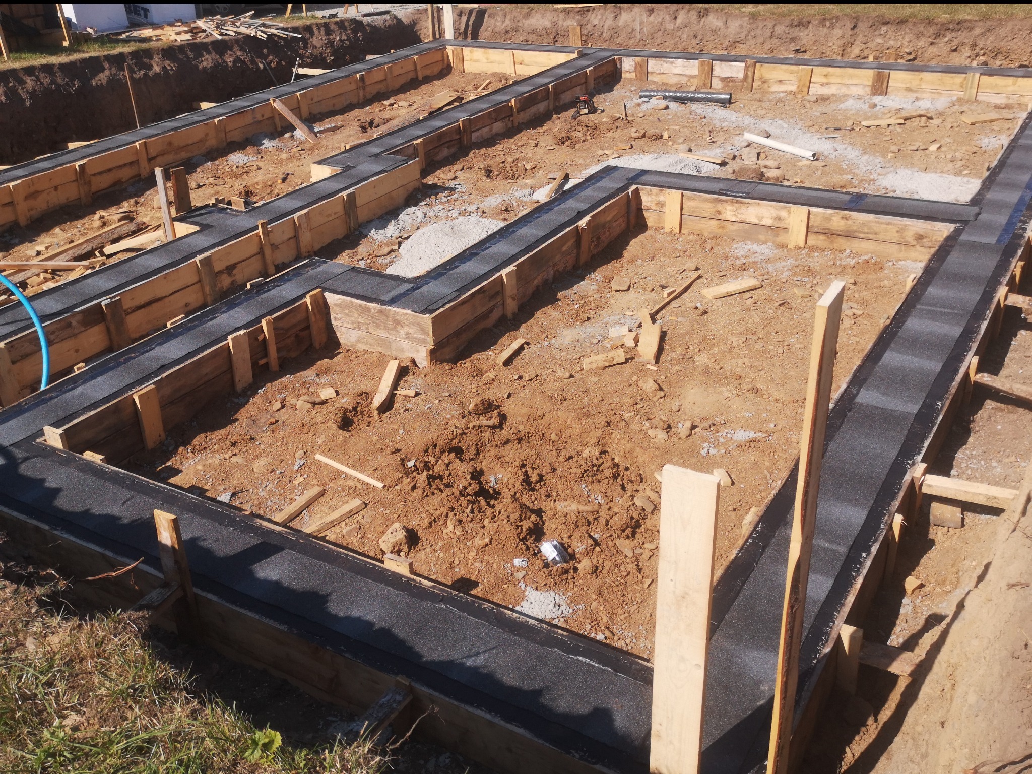 Szczegółowe ujęcie wykopu pod fundamenty z deskowaniem i izolacją przeciwwilgociową w postaci czarnej papy, widoczny fragment instalacji kanalizacyjnej.