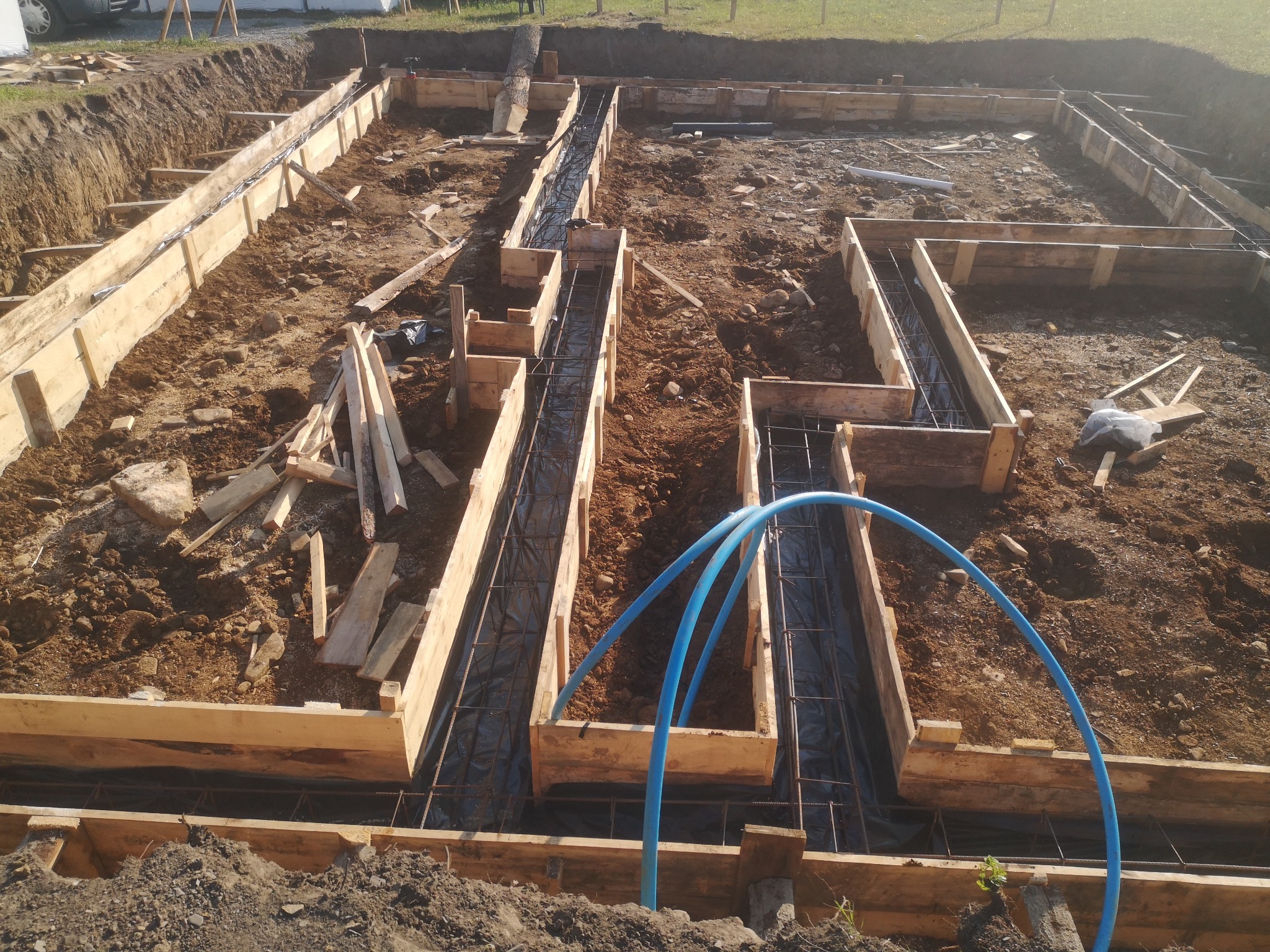 Przygotowanie szalunku i zbrojenia pod fundament domu jednorodzinnego, widoczne niebieskie rury instalacji podziemnej.