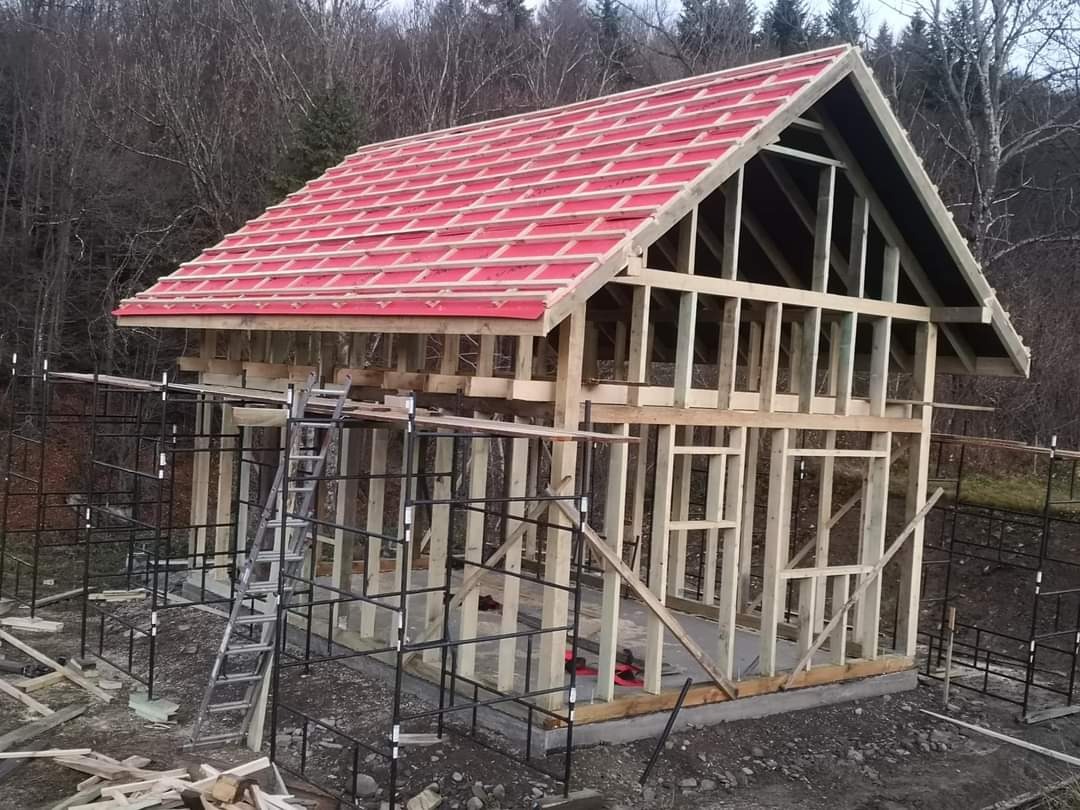 Wznoszona konstrukcja budynku z drewnianym szkieletem i częściowo ułożonym czerwonym dachem, widoczne rusztowania i drabina, tło stanowią drzewa.