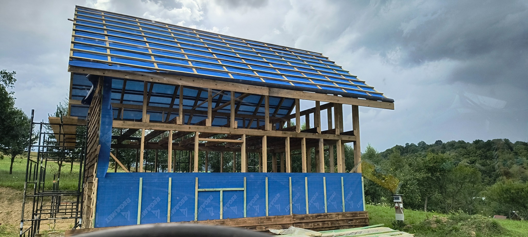 Konstrukcja drewniana domu w trakcie budowy, zabezpieczona niebieską folią, widoczne rusztowania i otaczająca zieleń. Ujęcie z dołu, pochmurne niebo w tle.