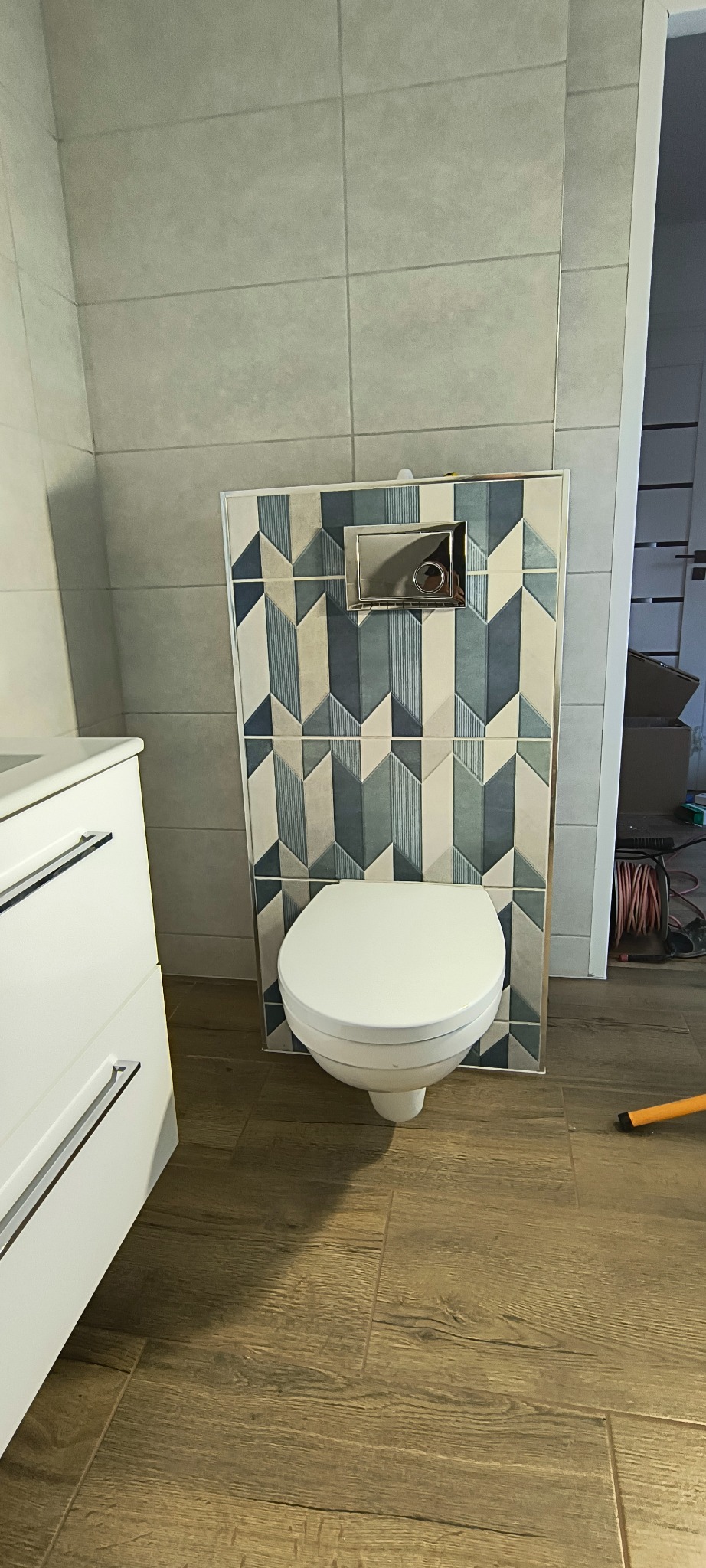 Nowoczesna toaleta podwieszana z geometrycznym wzorem płytek na ścianie, biała szafka łazienkowa z chromowanymi uchwytami, drewnopodobna podłoga.
