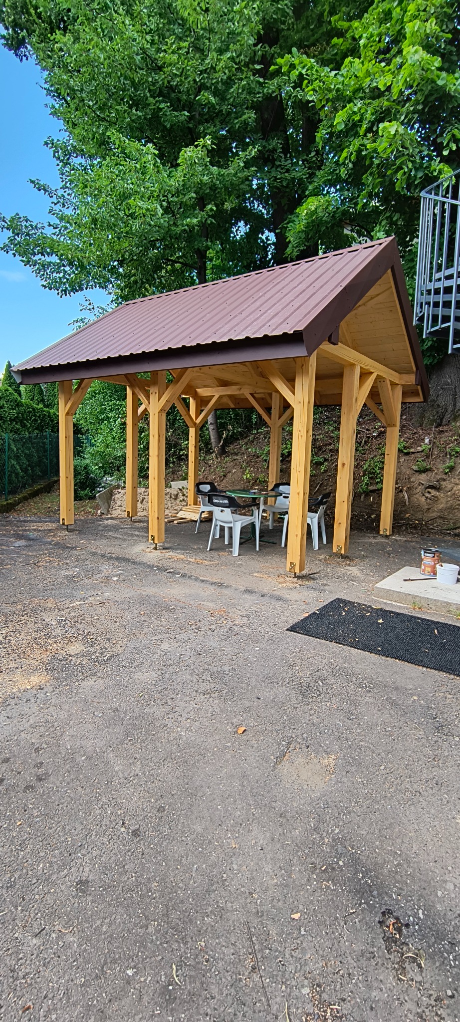 Drewniana altana z brązowym dachem, otoczona zielenią. Widoczne krzesła i stolik wewnątrz. Na ziemi leżą drobne ścinki drewna. Słoneczny dzień.