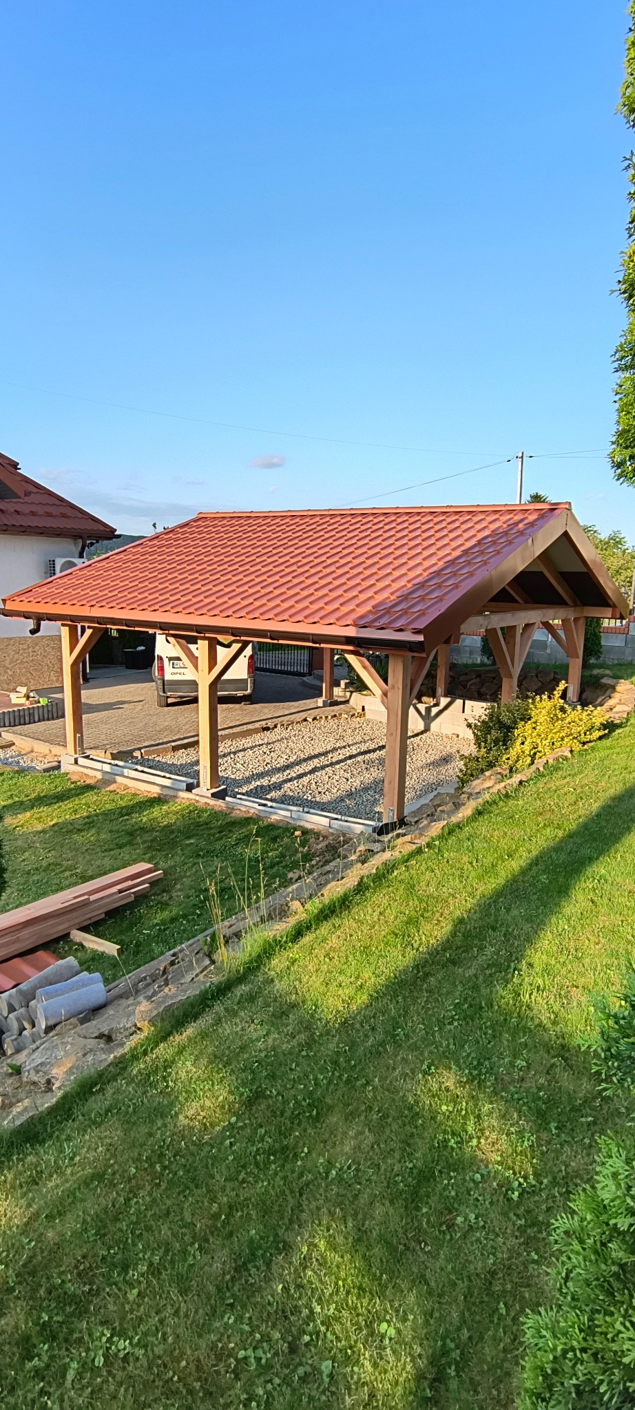 Drewniana wiata garażowa z czerwoną dachówką, otoczona zielonym trawnikiem i elementami krajobrazu. Częściowo widoczny dom i zaparkowany samochód w tle.