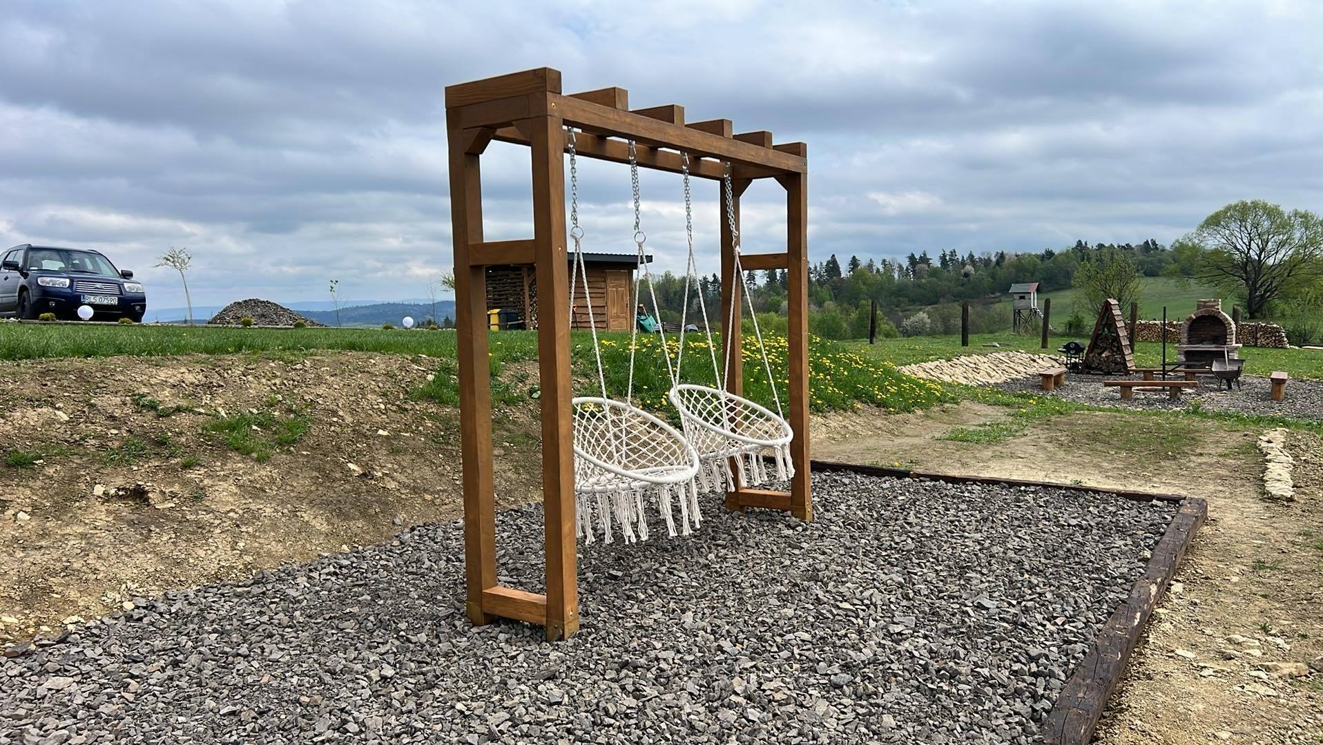 Drewniana konstrukcja z dwoma wiszącymi, plecionymi fotelami na żwirowym podłożu. W tle zieleń, drzewa i grill. W oddali widoczny samochód.