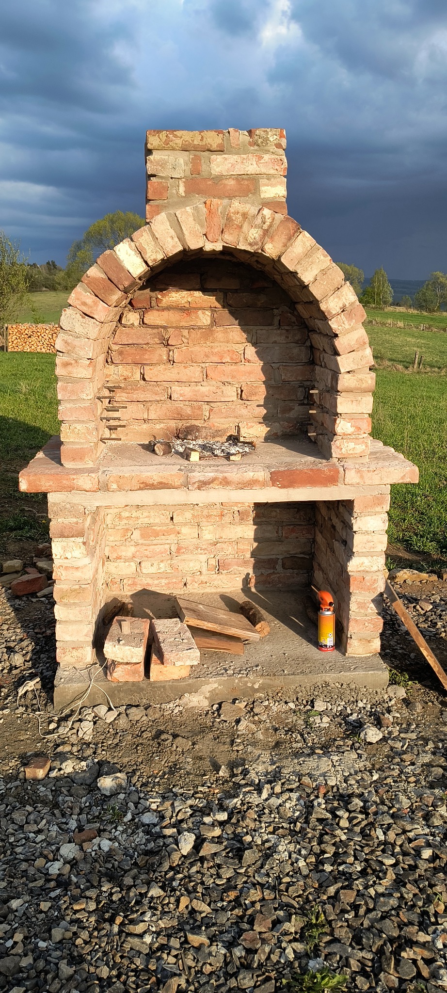 Murowany grill ogrodowy z czerwonej cegły w trakcie budowy, z widocznymi materiałami i narzędziami, na tle zielonej łąki i burzowego nieba.