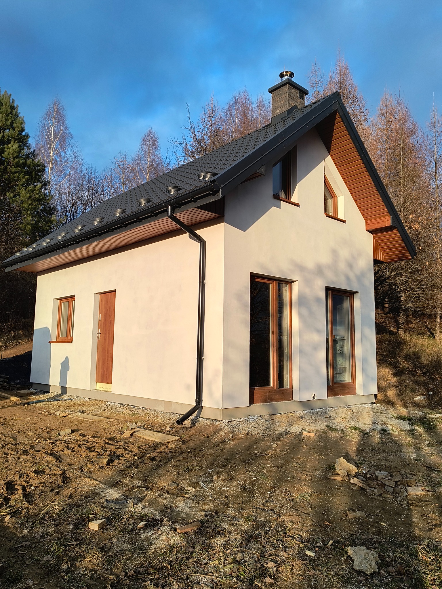 Niewielki, parterowy dom z ciemną dachówką i drewnianą stolarką okienną, otoczony naturalnym krajobrazem w jesiennej scenerii. Widok z boku, w słoneczny dzień.