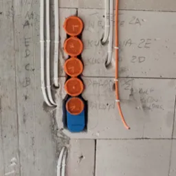Ściana z betonu komórkowego z pionowo ułożonymi przewodami elektrycznymi w białych peszlach oraz pomarańczowymi puszkami instalacyjnymi i niebieską puszką pod gniazdko.