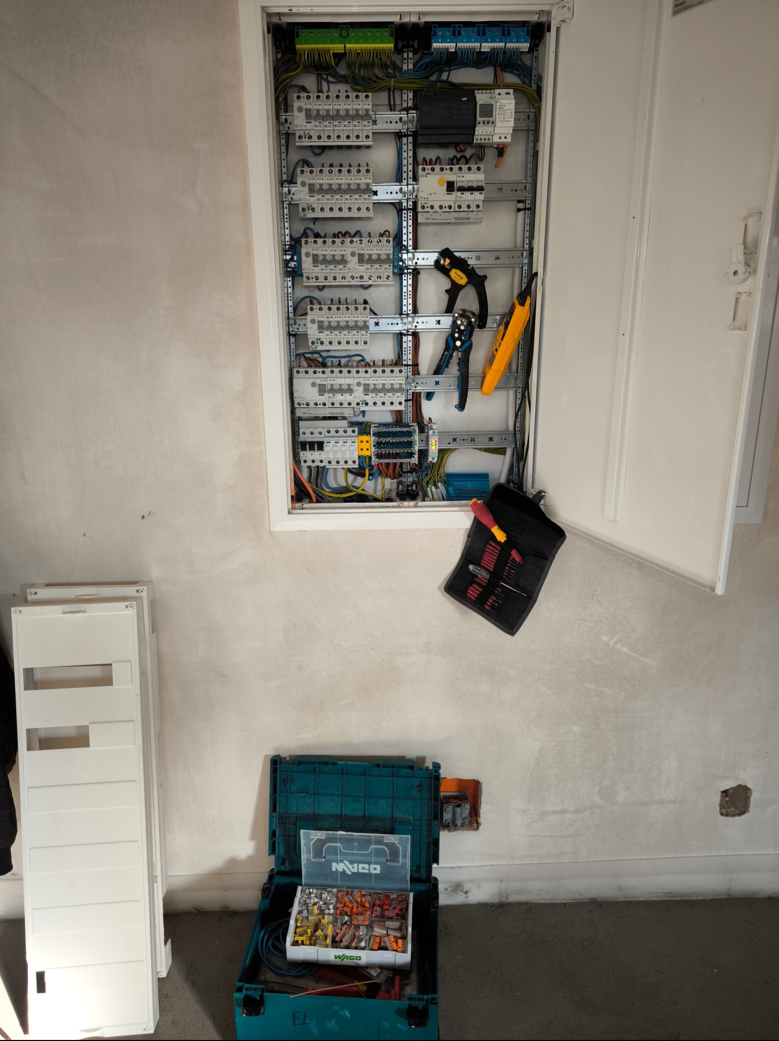 Rozdzielnia elektryczna w trakcie montażu w surowym wnętrzu. Widoczne narzędzia, skrzynka z elementami Wago i otwarte drzwi rozdzielnicy. Ściany przygotowane pod malowanie.
