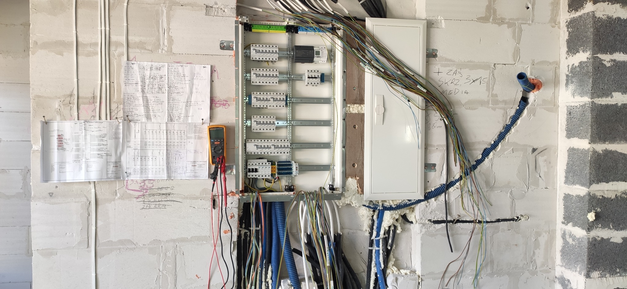 Skrzynka elektryczna z podłączonymi przewodami na ścianie z betonu komórkowego. Widoczne schematy i miernik elektryczny. Instalacja w trakcie montażu.