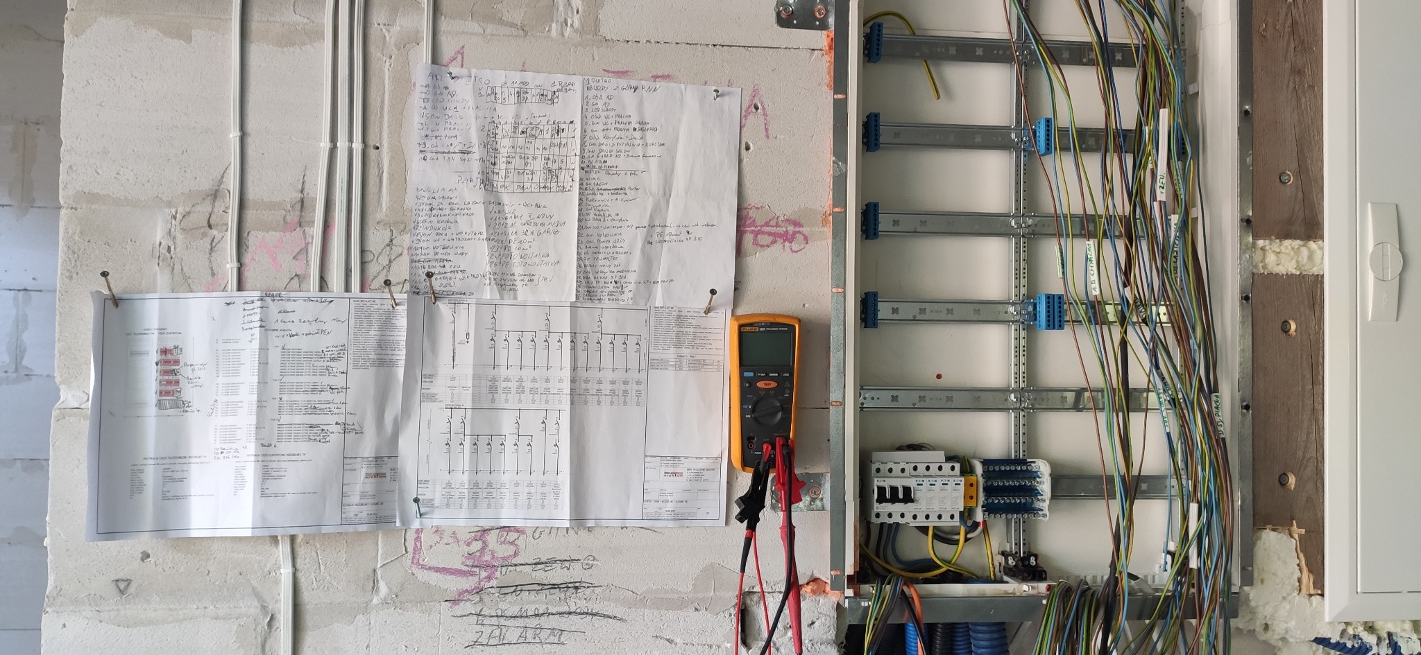 Rozdzielnica elektryczna w trakcie montażu, widoczne kable, schematy i miernik na ścianie z betonu komórkowego. Prace instalacyjne w surowym wnętrzu.