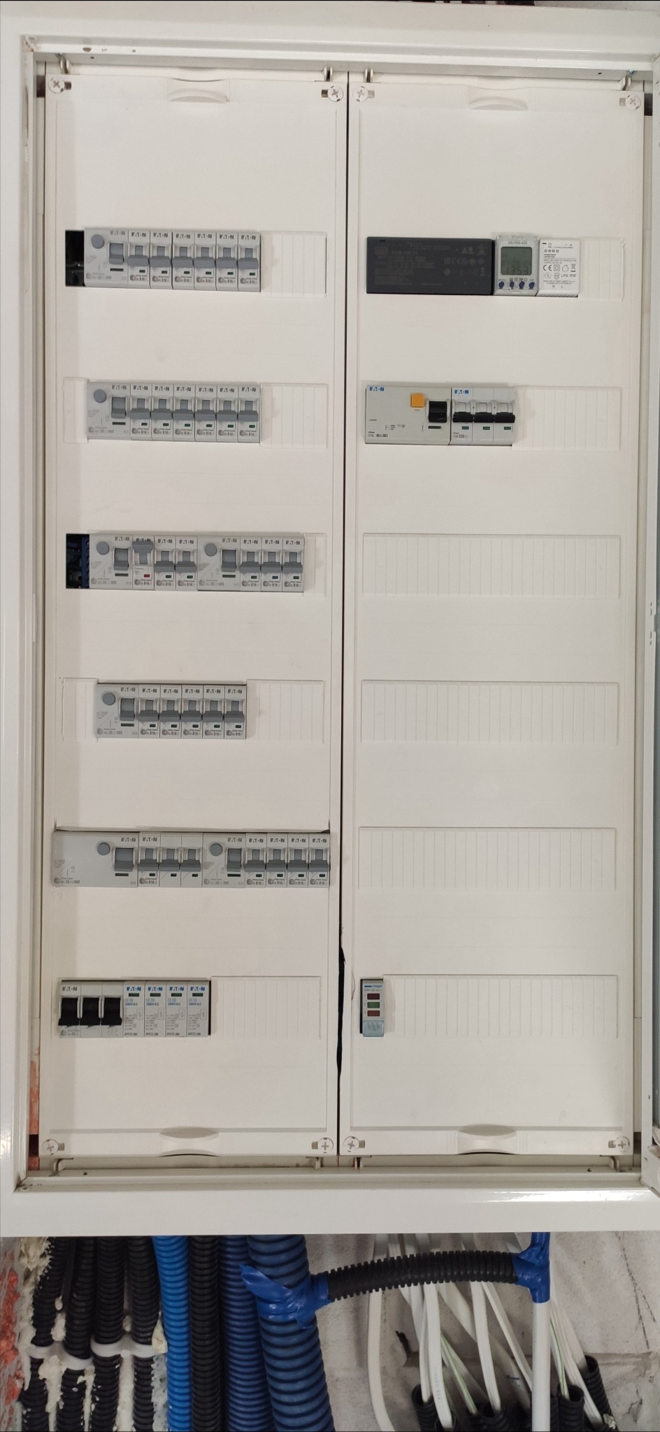 Rozdzielnica elektryczna Eaton z wyłącznikami nadprądowymi i różnicowoprądowymi. Widoczne niebieskie i czarne peszle z kablami. Biała obudowa wnęki.