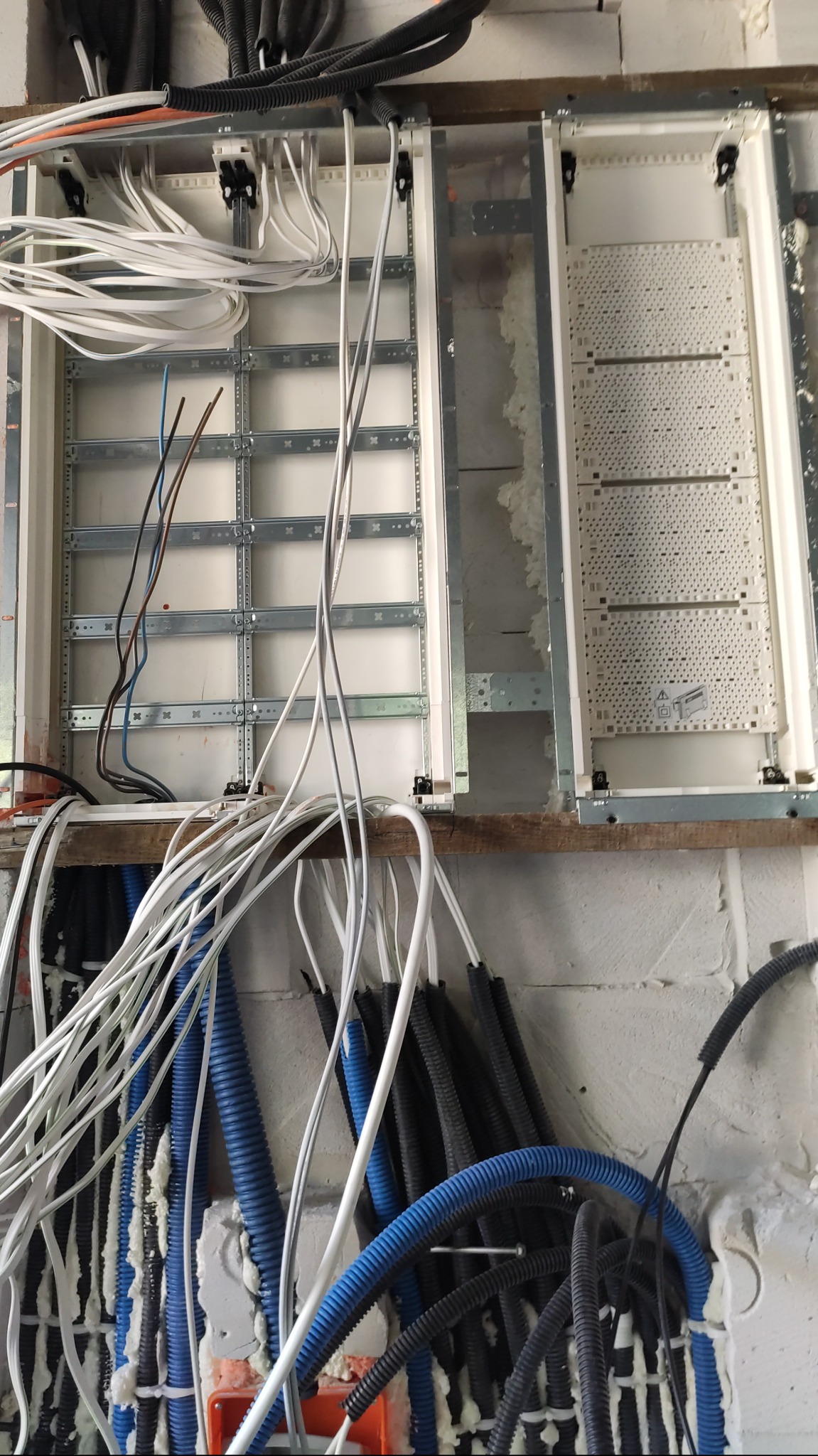 Rozdzielnia elektryczna w trakcie montażu: białe i niebieskie kable w peszlach, widoczne szyny montażowe, pianka montażowa uszczelniająca przejścia kablowe.
