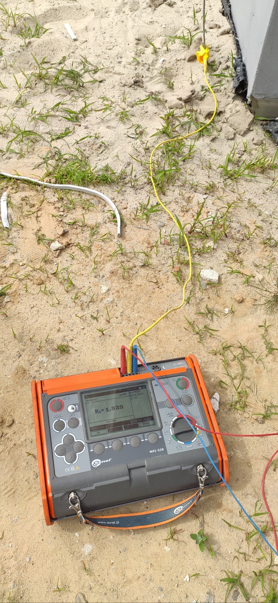 Miernik Sonel MPI-520 na piasku z podłączonymi przewodami pomiarowymi, wskazujący wynik pomiaru rezystancji uziemienia 1,92 Ω. Widoczne fragmenty instalacji elektrycznej.