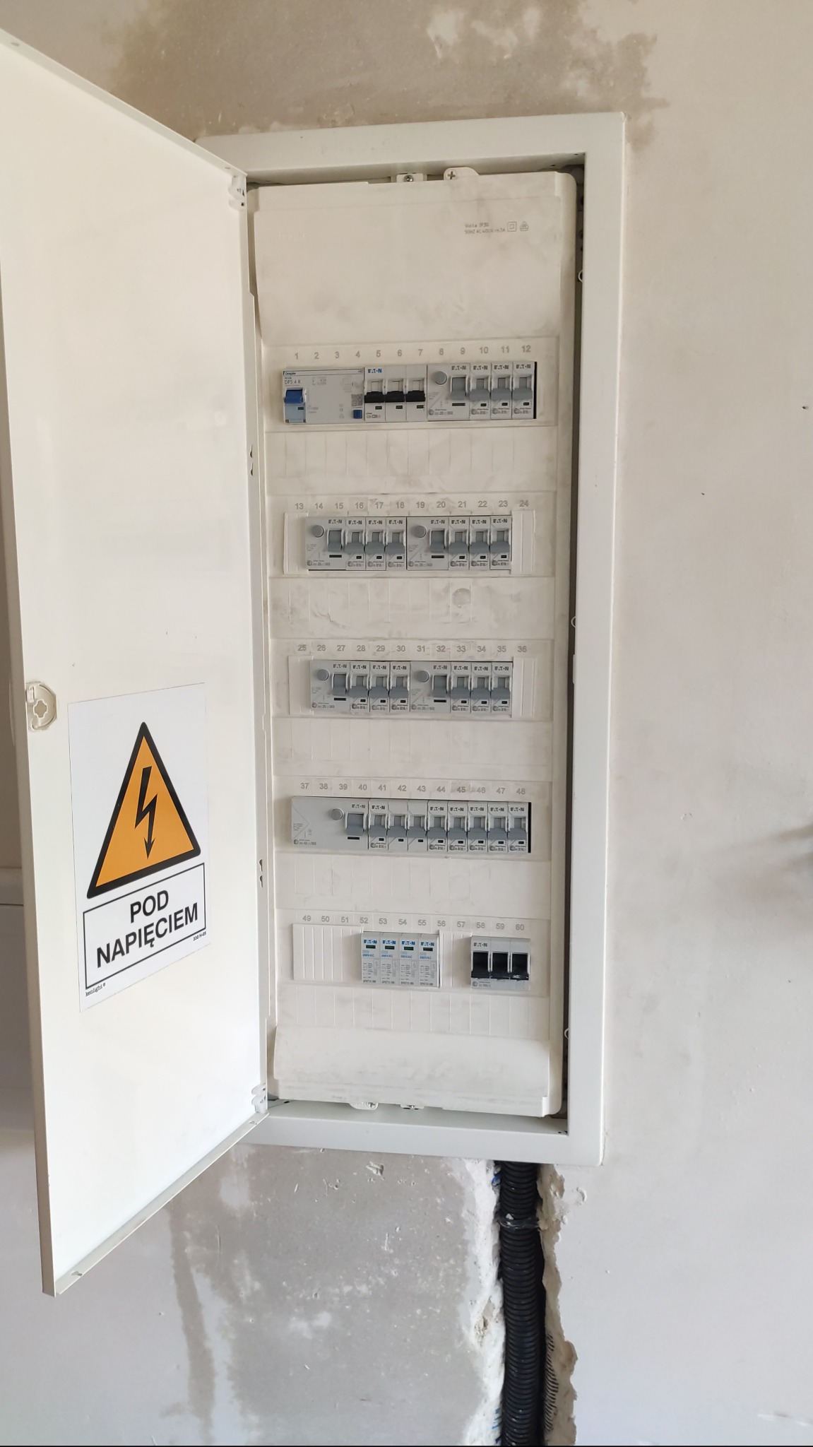 Otwarta skrzynka elektryczna z bezpiecznikami Eaton, widoczna tabliczka ostrzegawcza 'Pod napięciem', przewody w peszlu w ścianie. Biała obudowa, surowa ściana.