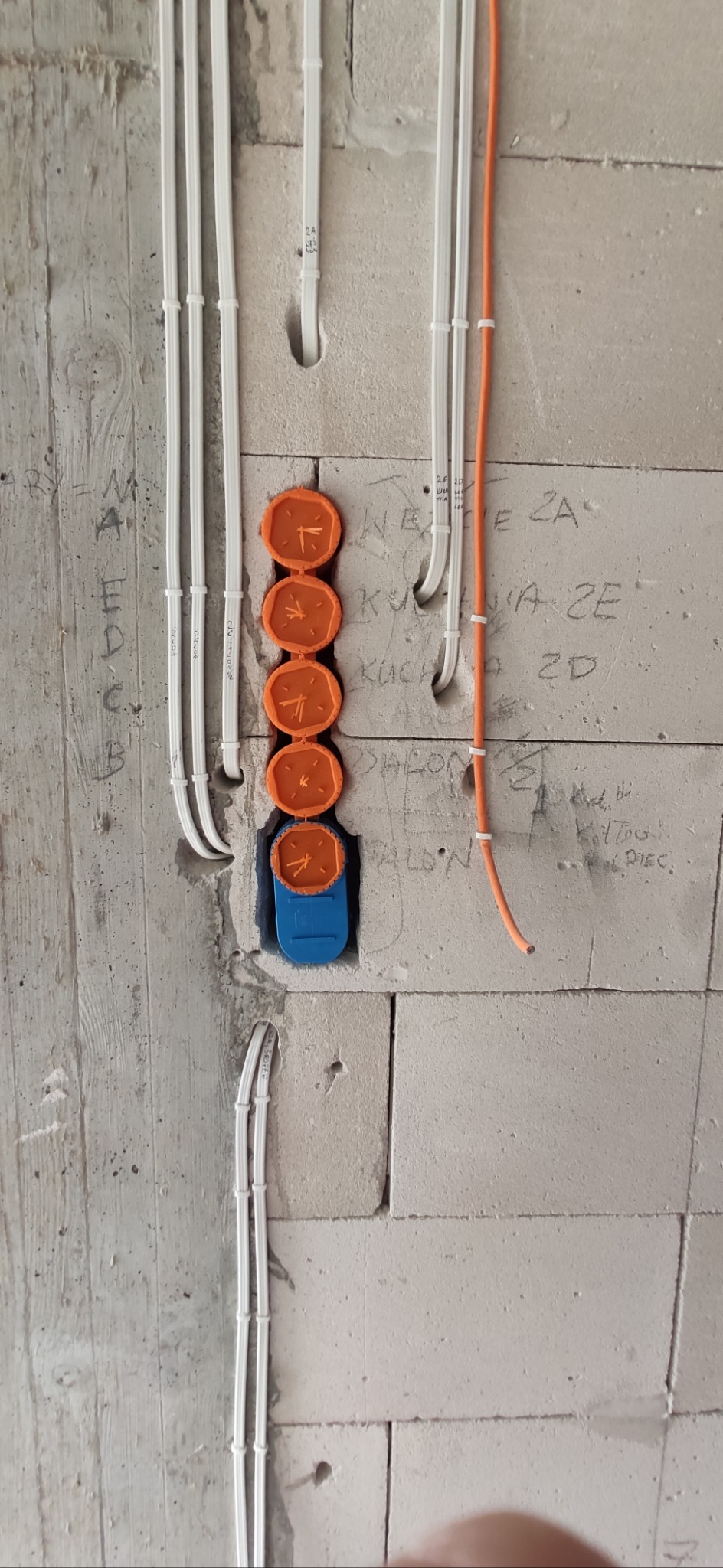 Ściana z betonu komórkowego z pionowo ułożonymi przewodami elektrycznymi w białych peszlach oraz pomarańczowymi puszkami instalacyjnymi i niebieską puszką pod gniazdko.