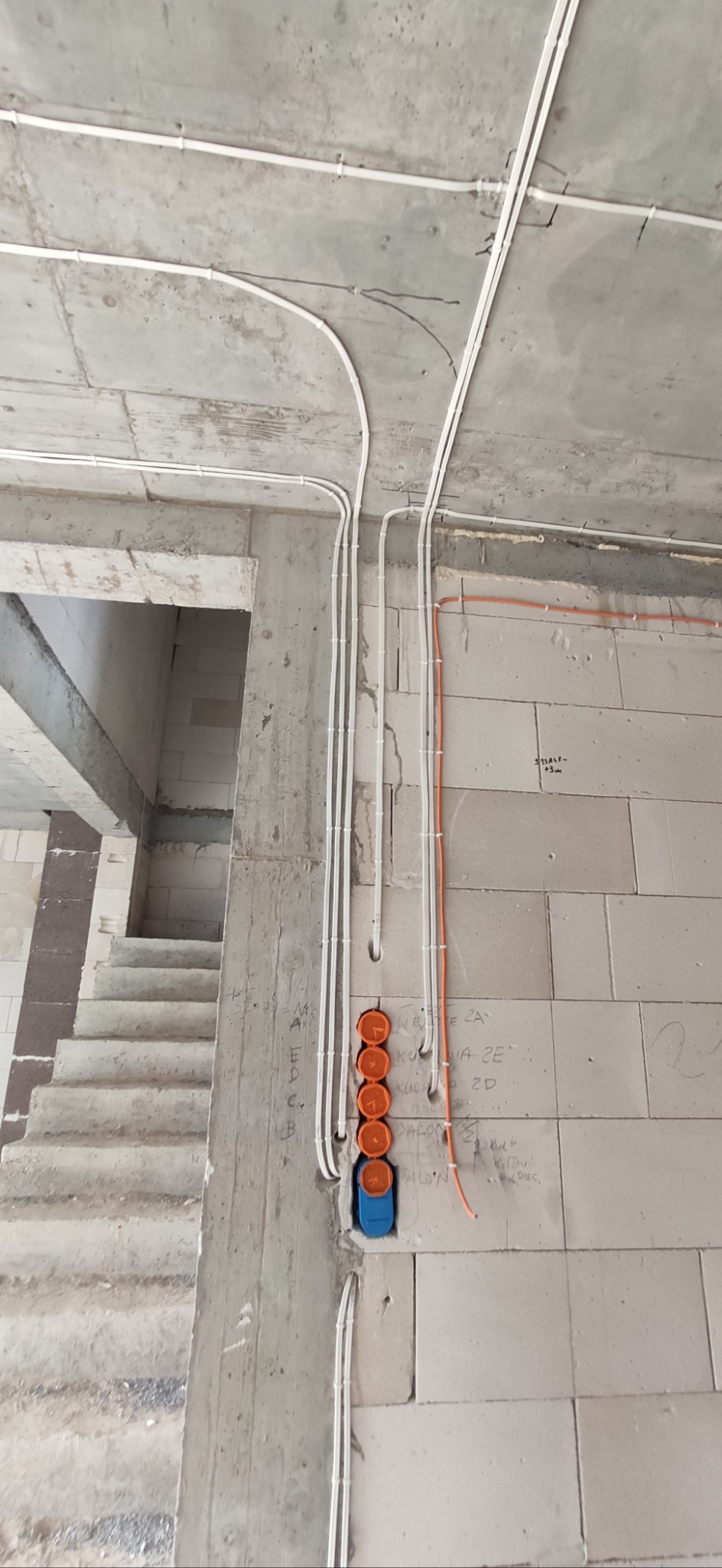 Surowy stan: Białe kable i pomarańczowe puszki elektryczne na ścianie z bloczków betonowych, widok na surowe schody w tle, instalacja w trakcie realizacji.