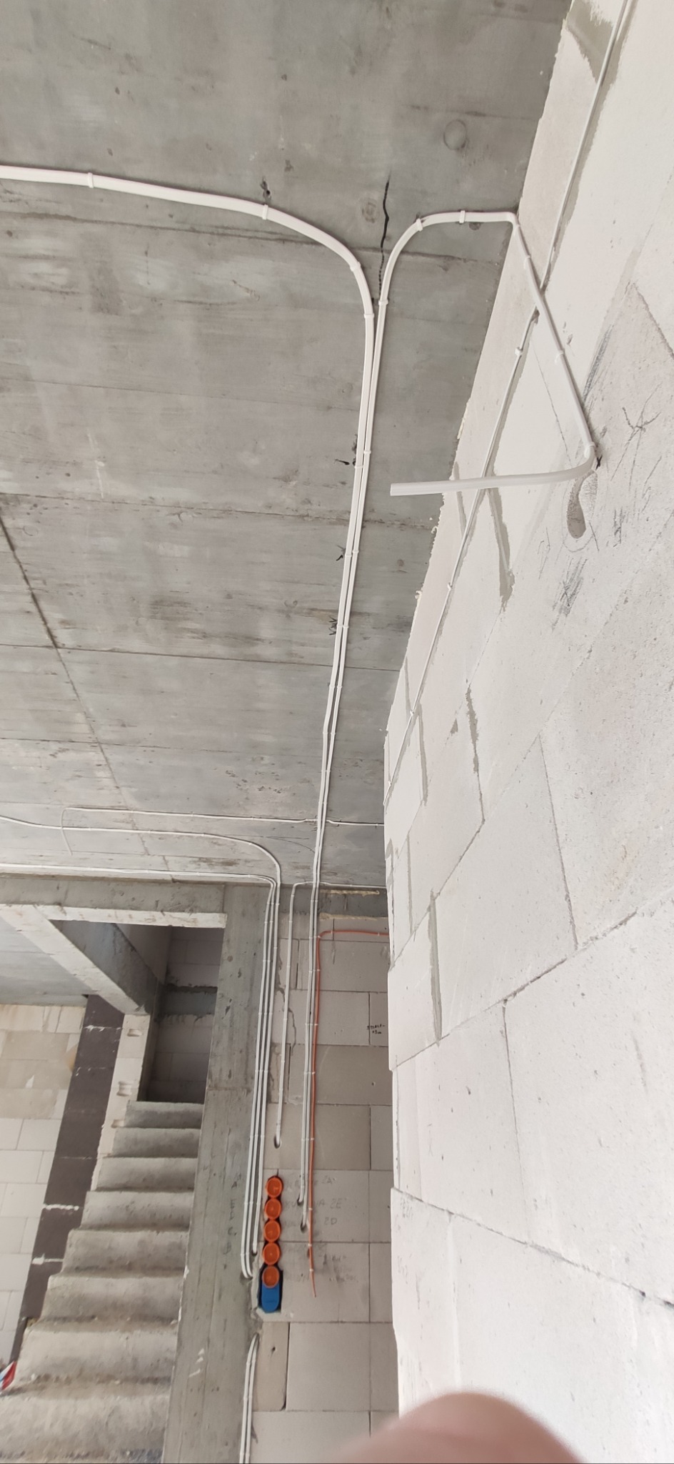 Białe przewody elektryczne ułożone na surowym, betonowym suficie i ścianie z białych bloków w trakcie budowy. Widoczne puszki instalacyjne i fragment schodów.