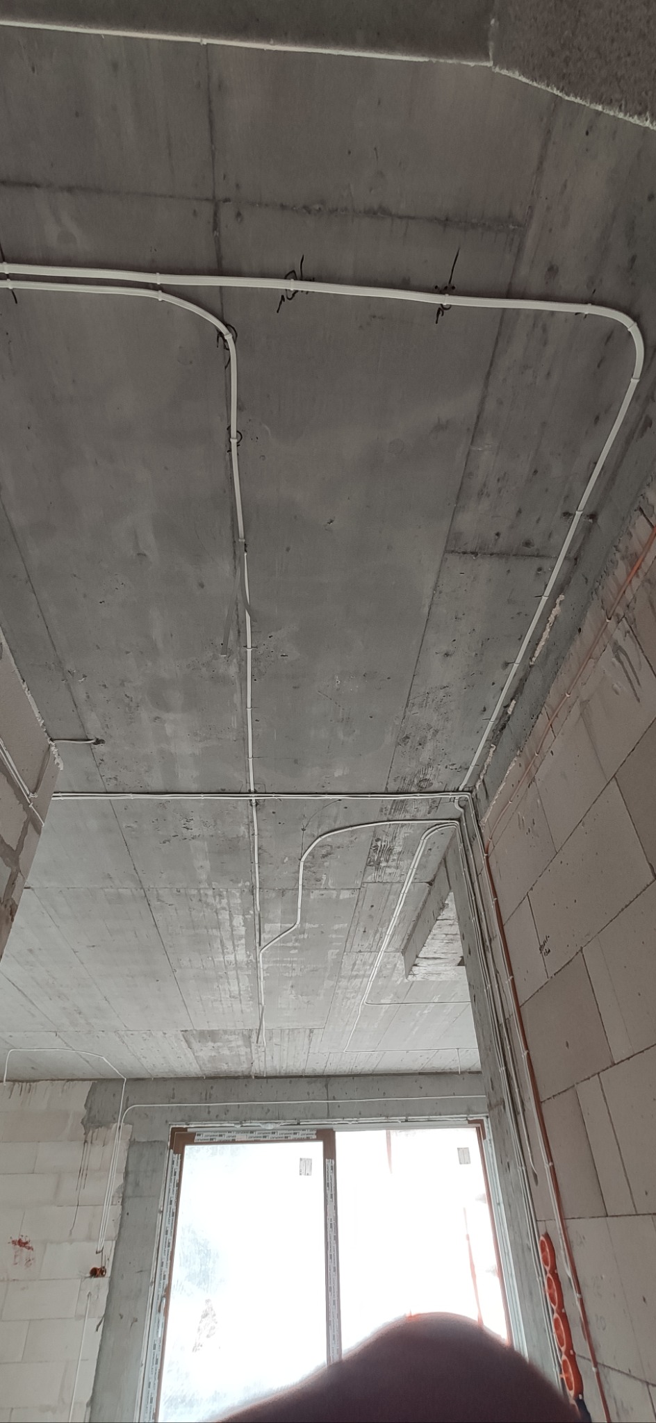 Białe kable elektryczne ułożone na surowym, betonowym suficie w nowym budynku. Instalacja prowadzona wzdłuż ścian i sufitu, widoczne mocowania kabli.