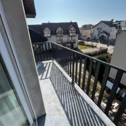 Ciemna, metalowa balustrada balkonowa rzuca wyraźne cienie na betonową podłogę balkonu w słoneczny dzień. W tle budynki mieszkalne.