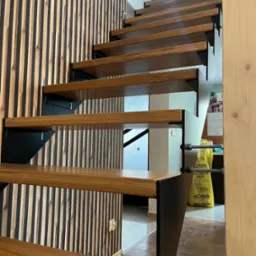 Drewniane schody metalowe z czarnymi wspornikami, widok z dołu na tle ściany z drewnianymi panelami, w tle żółte worki z tworzywa.